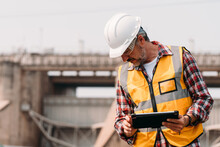 Portrait Of Power Engineer Wearing Safety Jacket And Hardhat With Tablet Working At Outdoor Field Site That Have Water Spillway  Of Hydro Power Dam Electrical Generator At The Background.