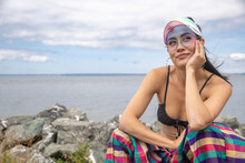Asian Woman Wearing A Visor And Bikini Top With Colorful Striped Pants Rests Her Hand Under Her Chin And Looks Up While Thinking. Ocean Water Is Behind Her As She Is Sitting On Shoreline Rocks. 