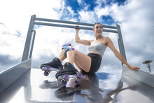 Woman Wearing Roller Skates Sitting On Top Of A Playground Park Slide. The Sky Is Blue With Puffy White Clouds. 