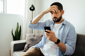 sad man checking smartphone sitting on a sofa at home