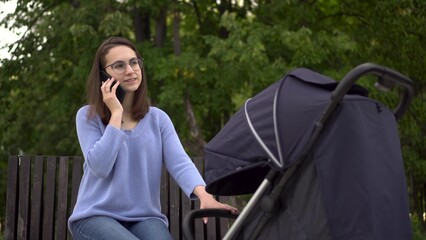 A young mother speaks on the phone and shakes a stroller with a child in the park. A woman with glasses speaks on a cell phone while sitting on a bench.