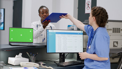 Nurse working with greenscreen template on laptop at facility registration desk. Using blank mockup display with isolated chroma key background and copy space on screen, reception counter.