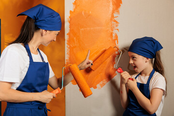 Canvas Print - Young people having fun painting at home, using orange paint color on roller paintbrush to redecorate apartment interior. Happy woman with little girl working on house renovation and decoration.