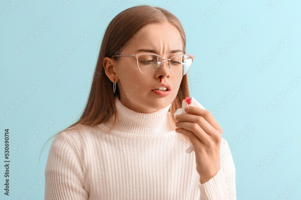 Young woman with nosebleed and tissue on blue background, closeup