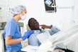Asian woman dentist listening to patient during consultation. African-american man pointing at tooth with problem.