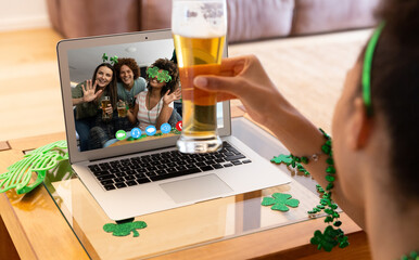 Poster - Mixed race woman holding beer having st patrick's day video call with friends on laptop at home