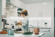 © simona - One woman cooking at home in the kitchen alone. Real single lifestyle for independent female people. Housewife preparing lunch for the family. View of lady using pot and prepare food indoor life.