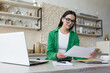 © Liubomir - Work remotely. A young beautiful businesswoman works from home. Sitting at home in kitchen working with laptop, documents and online calculator. Dressed in glasses and a green shirt.