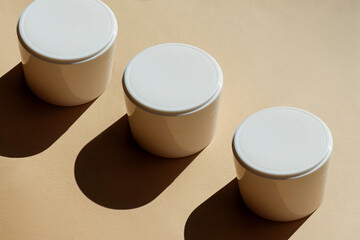 White jars of hand cream, faces on beige background in sunlight, with hard shadow. Unmarked containers for cosmetic products in close-up, on beige background