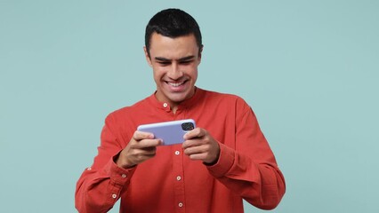 Wall Mural - Young fun gambling middle eastern man 20s he wear red shirt using play racing on mobile cell phone hold gadget smartphone for pc video games isolated on plain pastel light blue cyan background studio