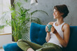 © ViDi Studio - Side view young woman wears white tank shirt drink tea coffee sit on blue sofa couch stay at home hotel flat rest relax spend free spare time in living room indoors grey wall. People lounge concept.