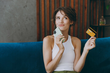 Wall Mural - Young pensive woman wear white tank shirt using mobile cell phone credit bank card shopping online booking tour sit on blue sofa stay home flat rest relax spend free spare time in living room indoors.