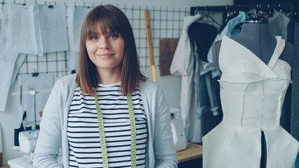 Wall Mural - Portrait of attractive young woman clothing designer with brown hair looking at camera. Smiling confident woman is standing beside dummy, light modern tailoring workshop in background.