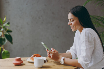 Sideways young smiling happy satisfied fun latin woman 30s wearing white shirt use mobile cell phone sit alone at table in coffee shop cafe restaurant indoors Freelance mobile office business concept