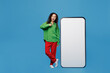 © ViDi Studio - Full body fun young woman of African American ethnicity in green shirt point finger on big huge blank screen mobile cell phone with workspace copy space mockup area isolated on plain blue background