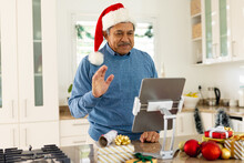 Happy Senior Biracial Man In Santa Hat Making Christmas Video Call On Tablet In Kitchen