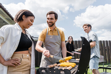 Smiling man in apron cooking corn on grill near bi-racial friend in backyard