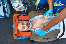 Ambulance Paramedic Using Defibrillator Performing Cardiopulmonary Resuscitation On Casualty. First Aid For Injured Mature Man Lying On Emergency Stretcher, Top View