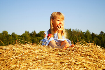 Little girl in blue dress sits on haystack. Eats red apple. Hot sunny day.