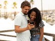 Diverse couple of happy heterosexual lovers looking at smartphone in a city on vacation
