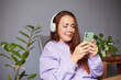 © leravalera89 - Cheerful caucasian female listening music or podcast and holding smartphone. A lot of green plants around. Red hair woman in violet sweater