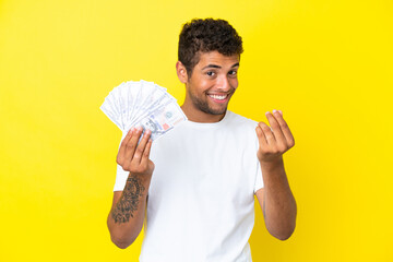 Young brazilian man taking a lot of money isolated on yellow background making money gesture