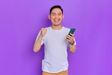 Excited Young Asian Man In White T-shirt Using Smartphone And Making Winner Gesture Isolated On Purple Background