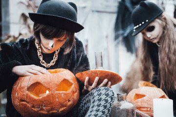 Beautiful scary little girl celebrating halloween. Terrifying black, white half-face makeup,witch costume, stylish image. Fun at children's party in barn on street. Hat,jacket, pumpkin jack-o-lantern