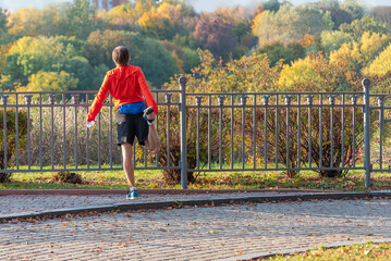 Wall Mural - Young caucasian male in bright sportswear performs physical exercises near the fence of autumn park. Fitness and healthy lifestyle in an urban environment.
