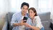 © tonefotografia - Young smiling asian couple using smartphone in living room at home