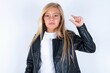 beautiful caucasian blonde little girl wearing biker jacket and glasses over white background purses lip and gestures with hand, shows something very little.