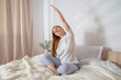 © TSViPhoto - Woman doing exercises in bed at home in the morning. Yoga pose side body stretch. Lady with long hair in pajamas. Girl stretches her muscles on a clean white bedding with a cozy blanket