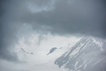  Atmospheric mountain landscape with fuzzy silhouettes of rocks on glacier in low clouds during rain. Dramatic view to large glacier tongue in snow mountains blurred in rain haze in gray low clouds.