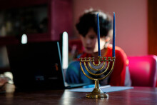 A Boy Sits At Table On A Computer, A Menorah In Foreground