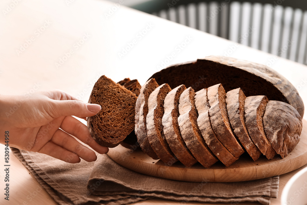 Woman taking slice of fresh bread from board in kitchen, closeup