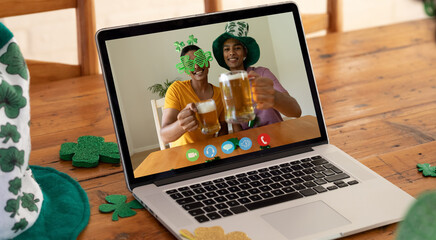 Webcam view of two african american men holding beers on video call on laptop on wooden table