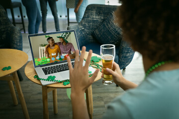 Wall Mural - Mixed race man holding beer at bar making st patrick's day video call waving to couple on laptop