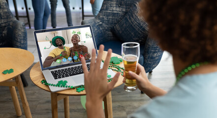 Wall Mural - Mixed race man holding beer at bar making st patrick's day video call waving to couple on laptop