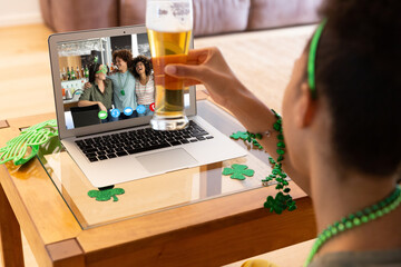 Canvas Print - Mixed race woman holding beer having st patrick's day video call with friends on laptop at home