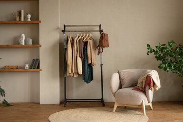 Rail rack with a capsule wardrobe in the living room against a beige wall. Trendy outfits in autumn colors: burgundy, beige, khaki, green, brown.