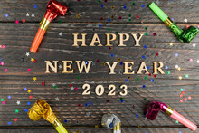 Happy New Year 2023 Wooden Letters On Wooden Background Surrounded Party Multicolored Whistles. Festive Greeting Card For New Year Holidays