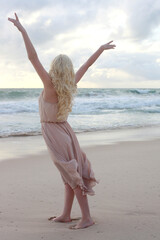 Canvas Print - full length portrait of beautiful young woman with long hair wearing flowing dress, standing pose walking away from the camera.  ocean beach background with sunset lighting.