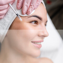 Wall Mural - Close up of hands of young cosmetologist injecting botox in female face. She is standing and smiling. The woman is closed her eyes with relaxation