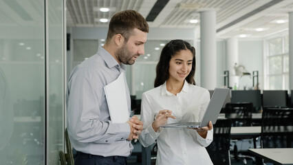 Positive friendly colleagues chatting in office discussing project details young female employee consults with male mentor shows presentation on laptop girl manager communicates co-worker in workplace