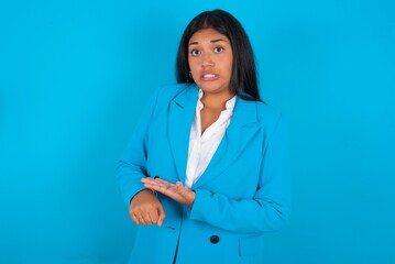 Wall Mural - Young latin woman wearing  blue blazer blue background In hurry pointing to watch time, impatience, upset and angry for deadline delay.