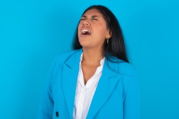 Wall Mural - Young latin woman wearing  blue blazer blue background angry and mad screaming frustrated and furious, shouting with anger. Rage and aggressive concept.