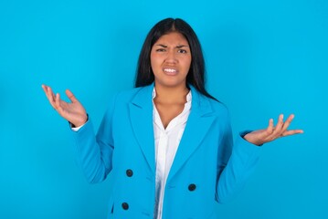 Wall Mural - Indignant Young latin woman wearing  blue blazer blue background gestures in bewilderment, frowns face with dissatisfaction.