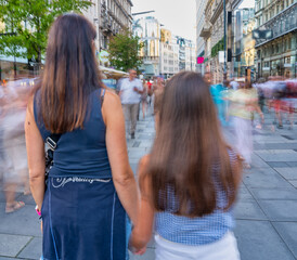 Sticker - Back view of a woman and her young daughter walking along a city street with blurred movement of tourists. Tourism and holiday concept