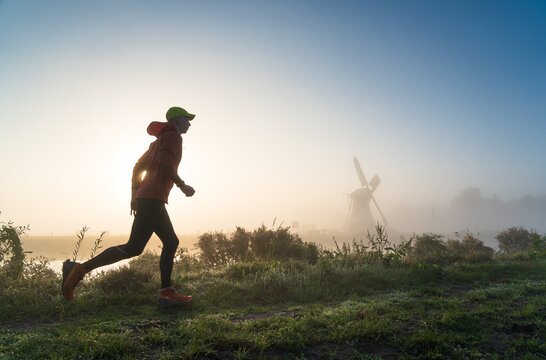 Fototapete -  - Man running in the foggy, Dutch countryside near a windmill during a tranquil sunrise. With motion blur.
