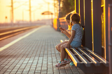 two little girls tourist sisters are sitting on the bench of the railway station and watching the phone while waiting for the electric train in the evening in the rays of the setting sun